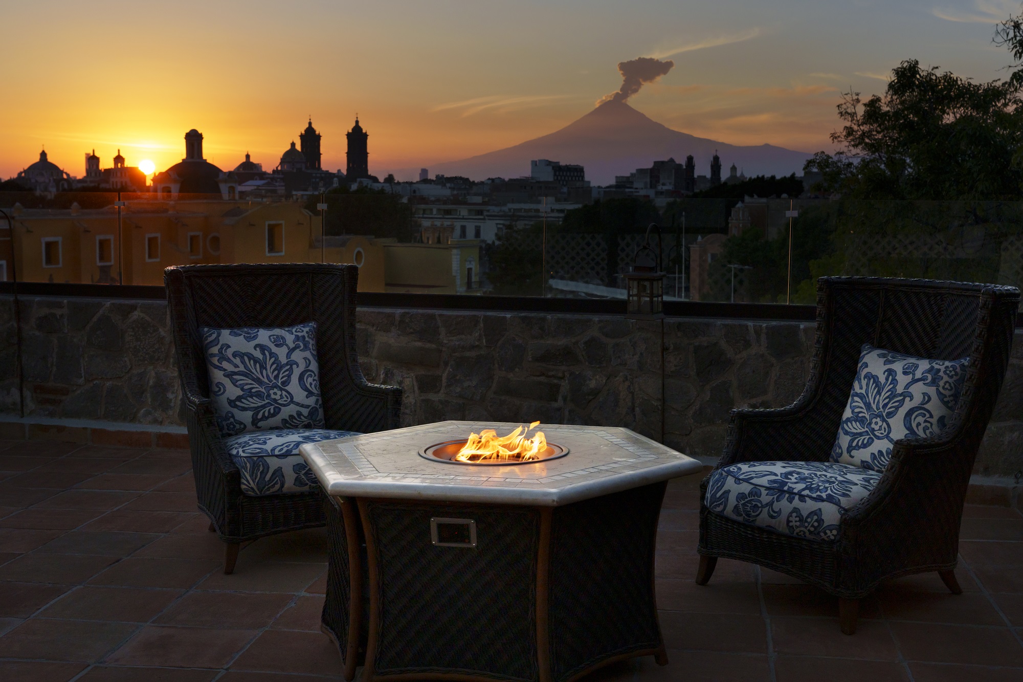 Patio chairs encircle a fire pit at Banyan Tree Puebla, with a sunset city skyline backdrop.